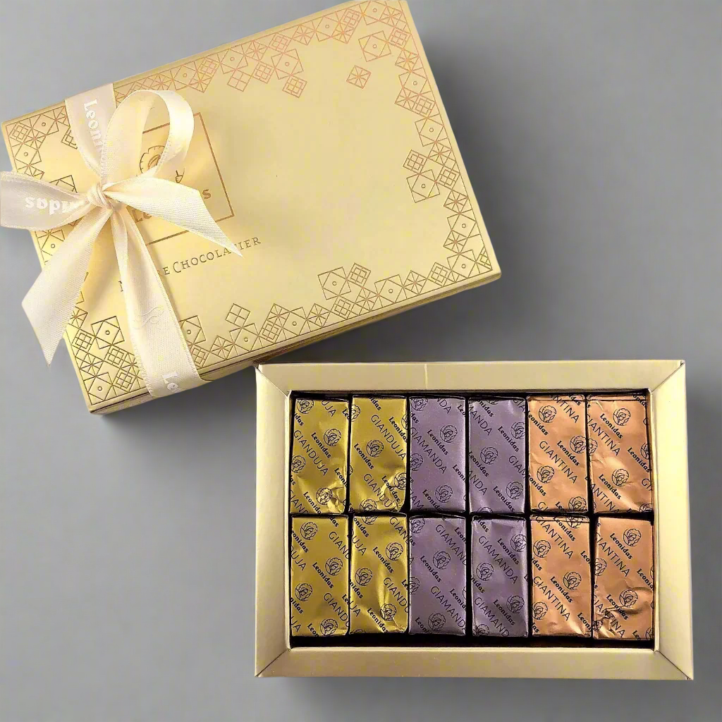 A gold box of Gianduja chocolates with a variety of flavors, including original, with Speculoos biscuit, with almond, and with nougat. The box is adorned with a decorative ribbon and placed on a wooden surface with flowers scattered around.